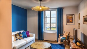 Een moderne woonkamer met een witte bank, kleurrijke kussens, een rond hockertje, een houten salontafel, een televisie en een groot raam met blauwe gordijnen.