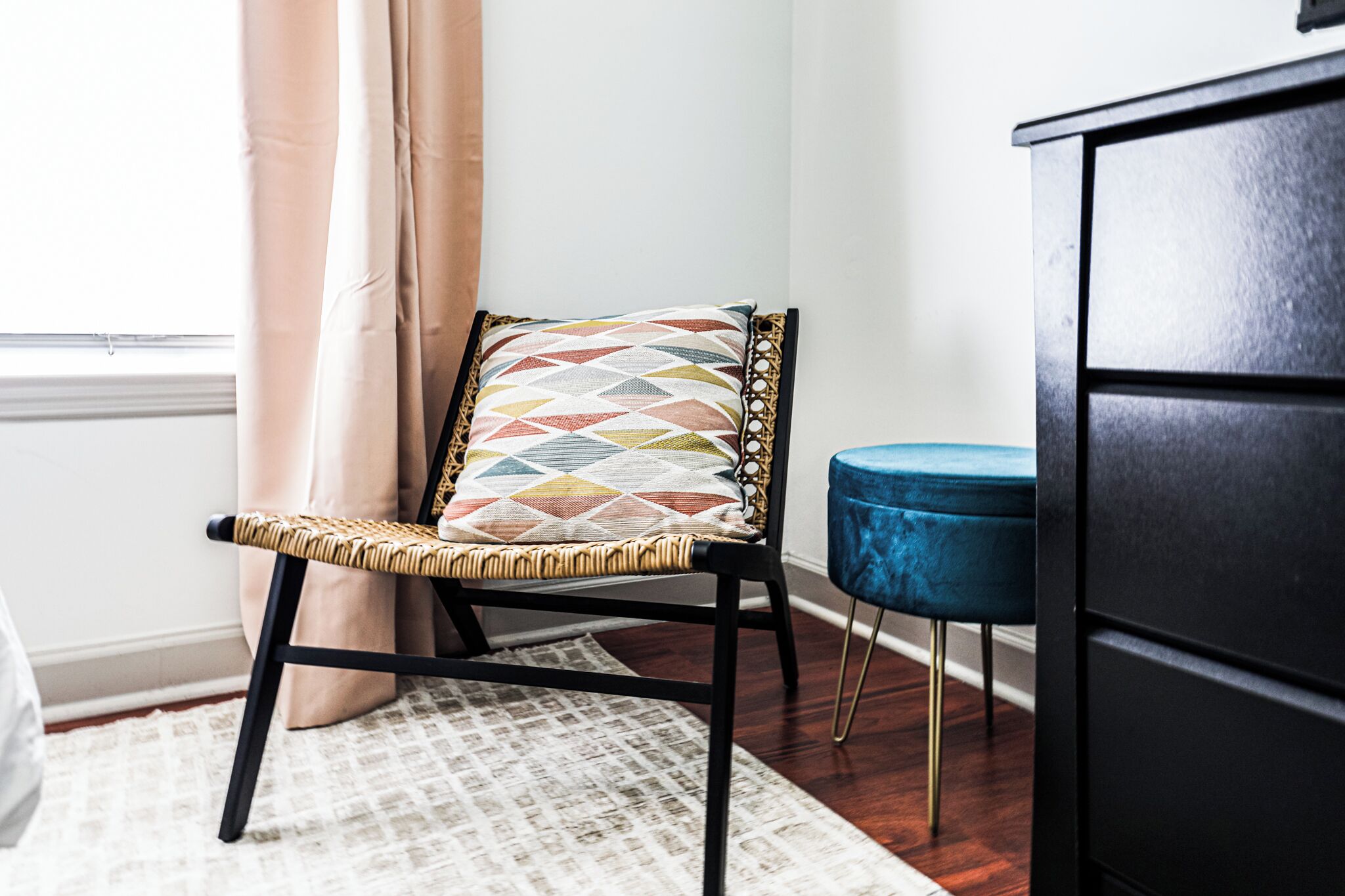 A chair with a geometric patterned cushion, a blue ottoman, and a black cabinet in a room with a patterned rug and a window with curtains.