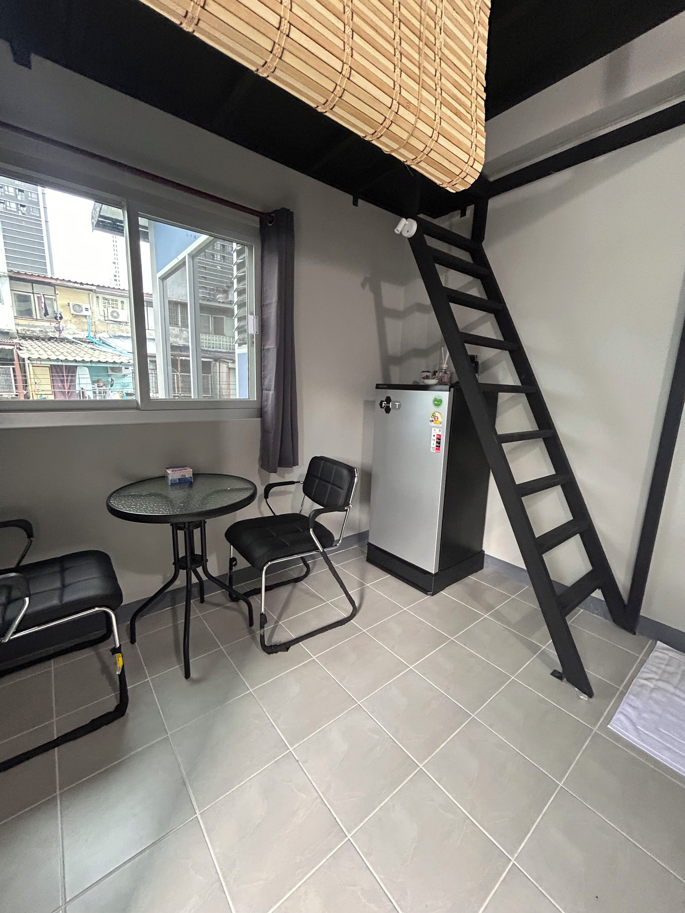 A small room with a table and two chairs, a refrigerator, a window with blinds, and a black ladder against the wall.