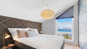 A modern bedroom with a large bed, a headboard with a geometric pattern, bedside lamps, and a view of a snowy landscape through a large window. - Deluxe Villa by Paramount (Troms)