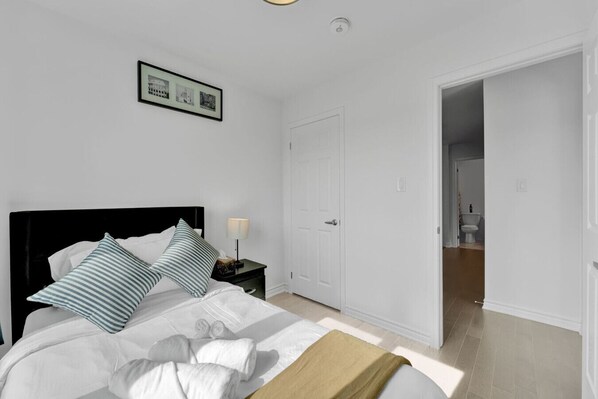 A bedroom with a bed, a nightstand, a lamp, a door, and a hallway leading to a bathroom.