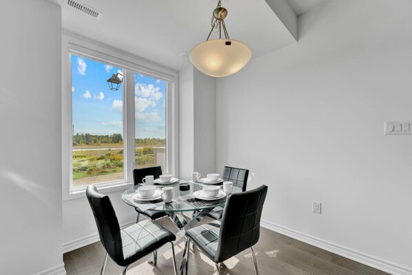 A dining area with a glass table and chairs, a view of a field through large windows, and a hanging light fixture.