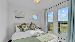Un dormitorio con una cama grande, dos almohadas decorativas, una mesita de noche con una lámpara y vistas a un balcón por una ventana amplia.