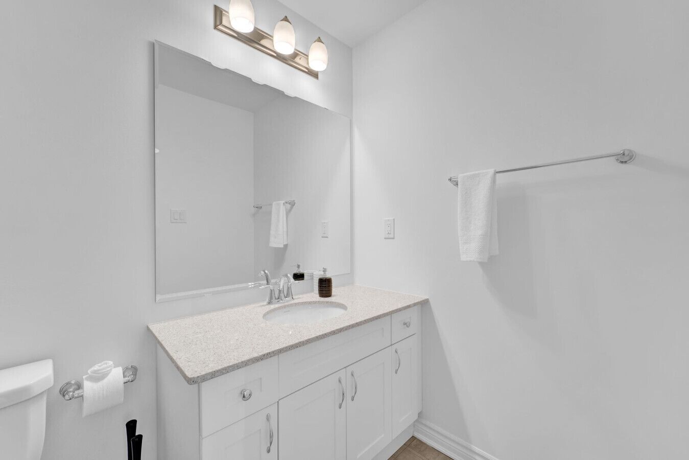 Un baño moderno con un lavabo, tocador y espejo blancos. Hay un perchero en la pared y un inodoro con un dispensador de toallas de papel.