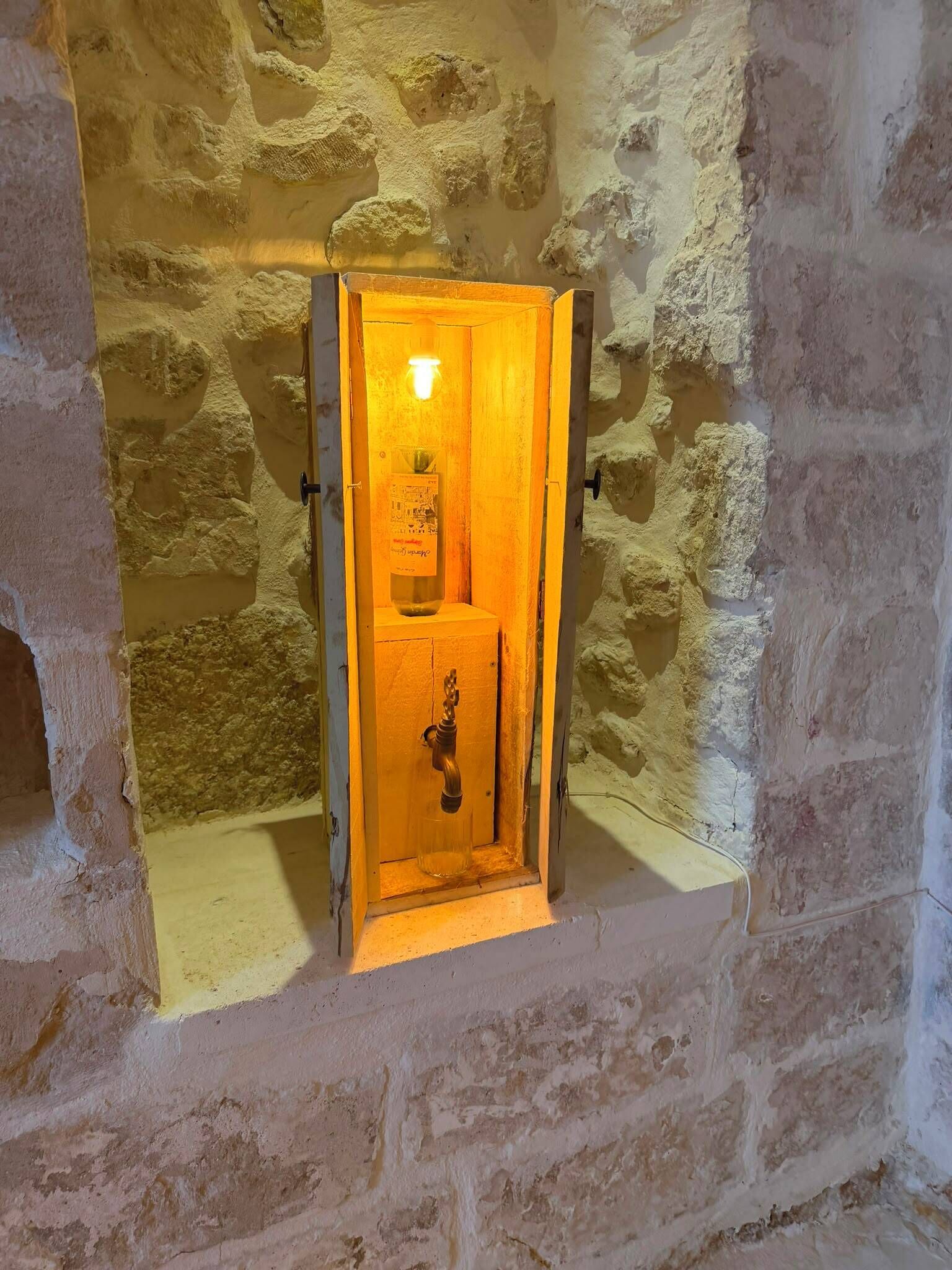 A stone-walled room with a wooden door, a light fixture, and a small shelf with a bottle.
