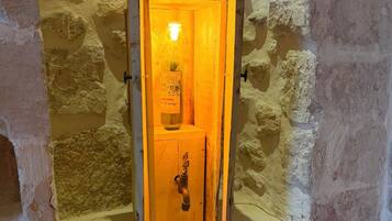 A stone-walled room with a wooden door, a light fixture, and a small shelf with a bottle.