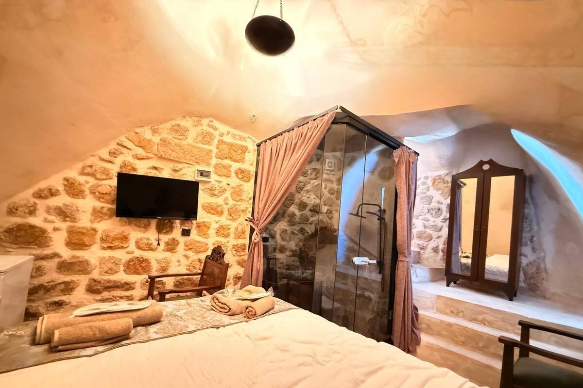 A cozy bedroom with a stone wall, a bed with rolled towels, a wooden chair, a TV, and a glass-enclosed shower area.