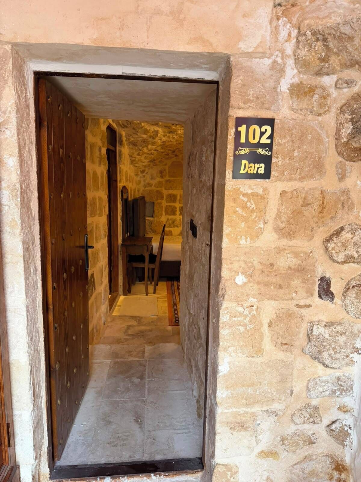 En korridor med stenmurar leder till ett rum med en trädörr. En skylt visar att det är rum nummer 102, och en stol syns i bakgrunden.