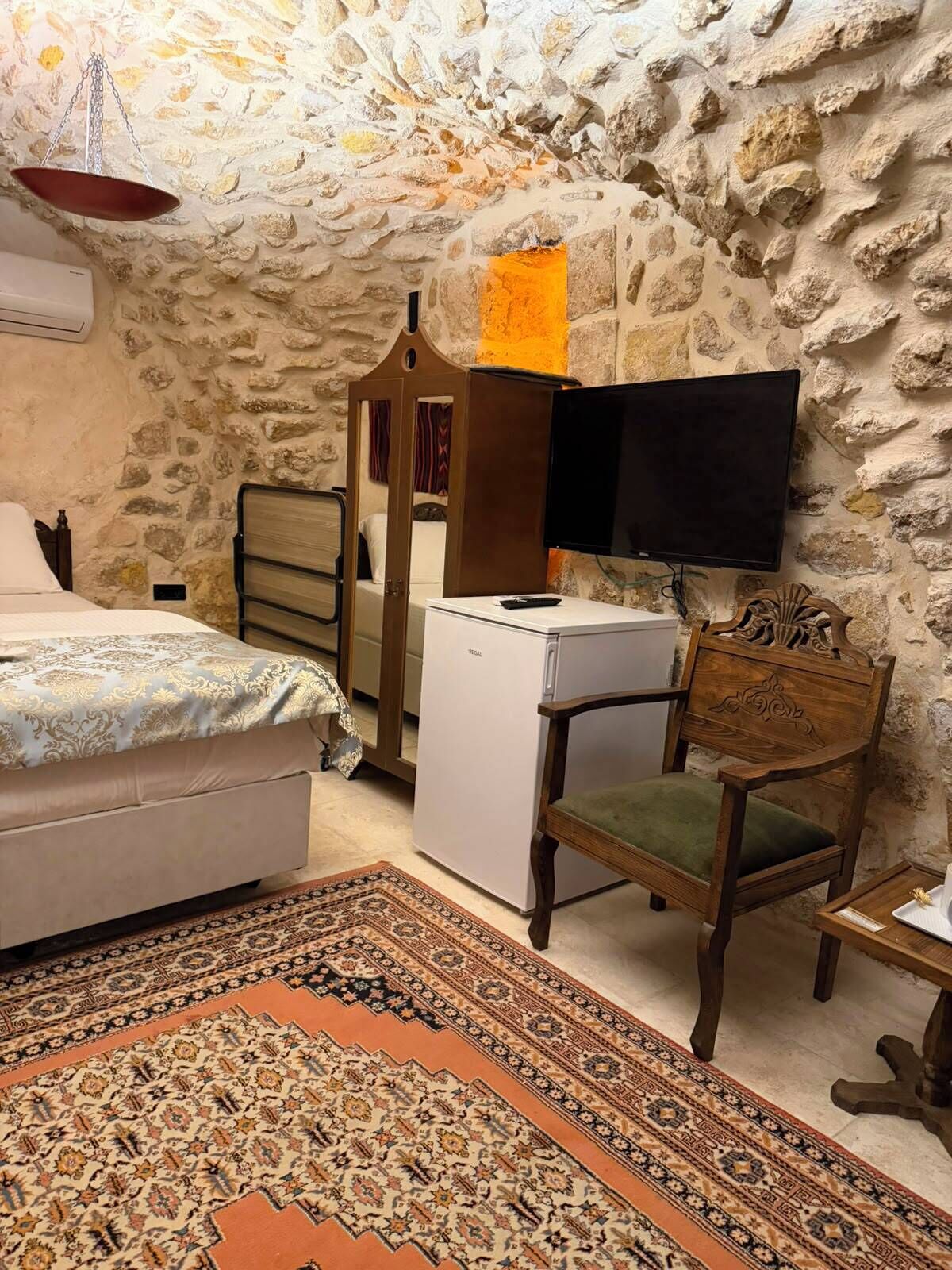 A bedroom with a stone wall, a bed, a desk with a television, a chair, and a small refrigerator.