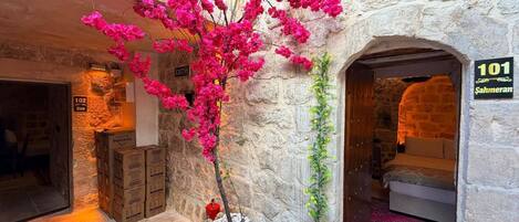 Interior de paredes de piedra con una planta de flores rosadas, un extintor y un cartel con el número de habitación.
