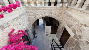 Un patio de paredes de piedra con un arco central, una escalera y flores rosadas a la izquierda.