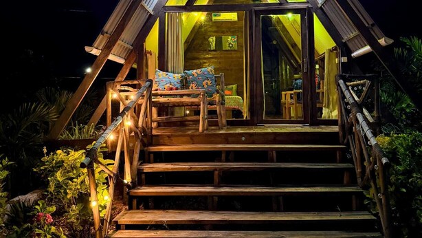 A triangular wooden structure with a staircase leading to an entrance, surrounded by plants. - leoglampingznz (Nungwi)
