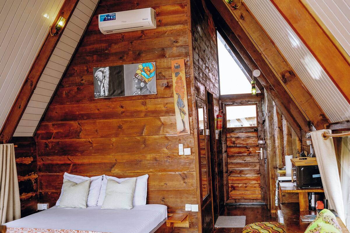 A cozy attic bedroom with a wooden sloped ceiling, a bed with a floral bedspread, a chair with a colorful cushion, and an air conditioning unit.