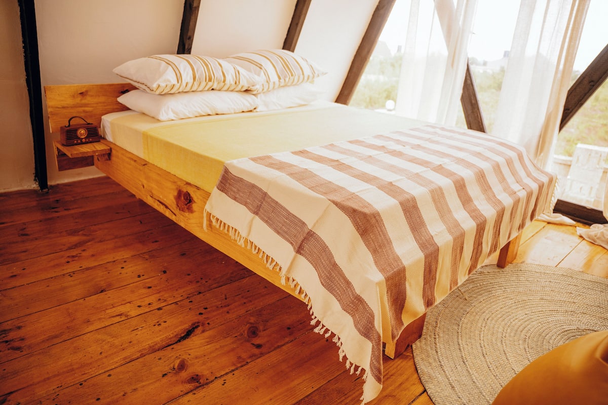 A wooden bed with a striped blanket, a radio, and a woven rug in a room with wooden floors and a window.