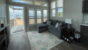 Een ruime woonkamer met een grijze modulaire bank, een vloerkleed met patroon en een plafondventilator. Grote ramen bieden uitzicht op het strand en de oceaan.