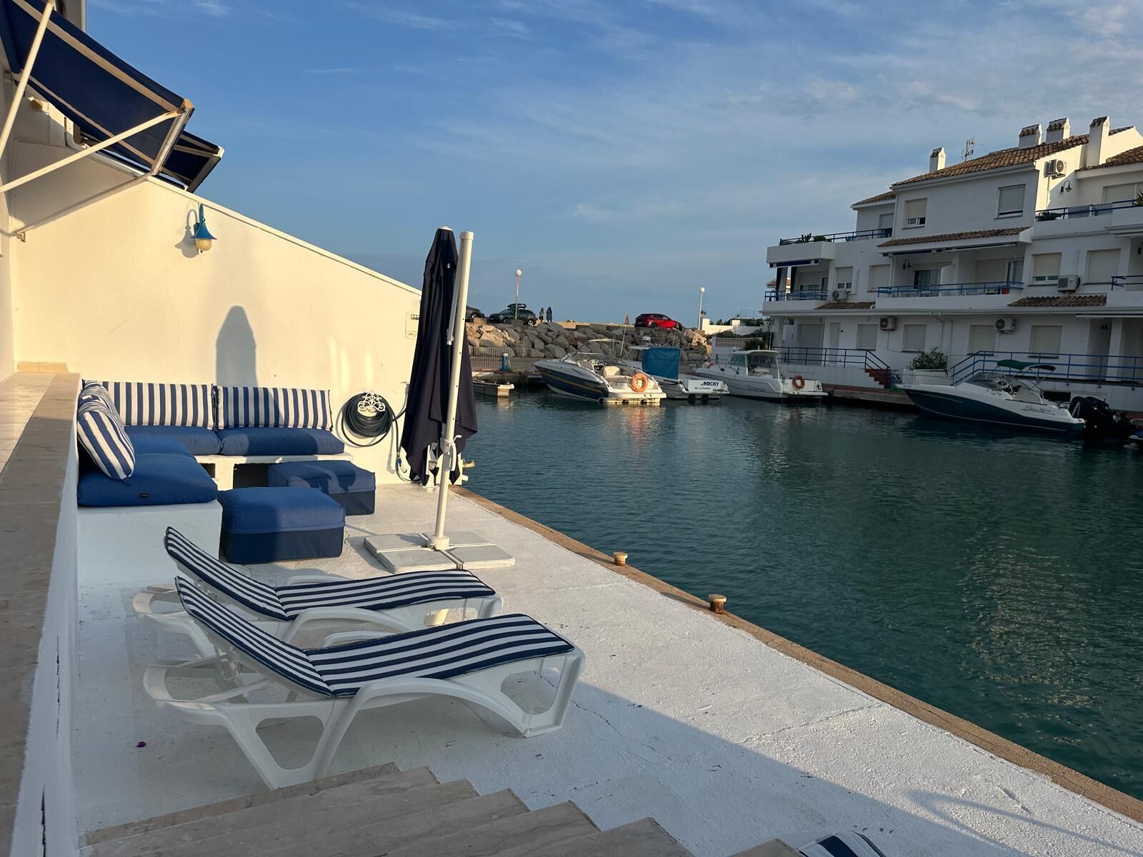 Un port de plaisance doté d’un espace détente avec des chaises longues à rayures et une table, offrant une vue sur une étendue d’eau calme où sont amarrés des bateaux.