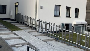 Un edificio moderno con techo plano, múltiples ventanas y un acceso pavimentado con escalones y barandilla.
