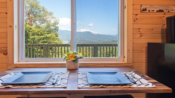 A wooden cabin with a dining table set for two, a window with a view of trees and mountains, and a sign on the wall.