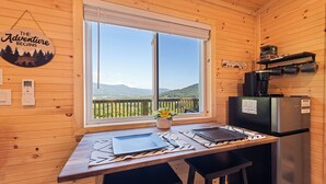 A wooden cabin interior with a dining table, a refrigerator, and a coffee machine. A window offers a view of a mountainous landscape.