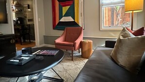 Una sala de estar moderna con un sofá de cuero negro, un sillón rojo, una mesa de centro con controles remotos, un cuadro abstracto de colores y una ventana con una lámpara.