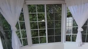 A bedroom with a bed, a window with curtains, and a view of greenery outside. - Ocho Rios Luxury Ocean View (Ocho Rios)