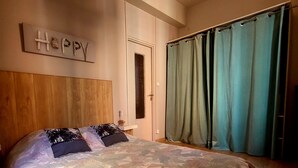 A bedroom with a floral bedspread, a wooden headboard, a 'Happy' sign, a window with blinds, and teal curtains. - Furnished stopover (Belfort)