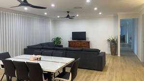 A modern living room with a dark-colored sofa, a wooden cabinet with a flat-screen TV, a dining table with chairs, and a ceiling fan. - Relaxing Acreage Retreat – Pool, Spa & Media Room (Karalee)