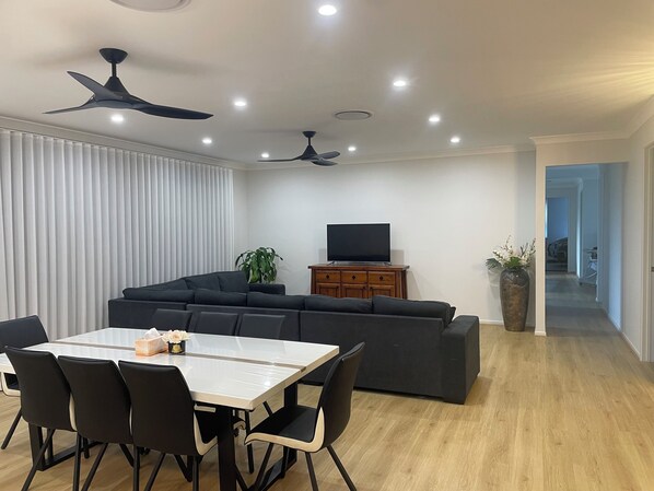 A modern living room with a dark-colored sofa, a wooden cabinet with a flat-screen TV, a dining table with chairs, and a ceiling fan. - Relaxing Acreage Retreat – Pool, Spa & Media Room (Karalee)