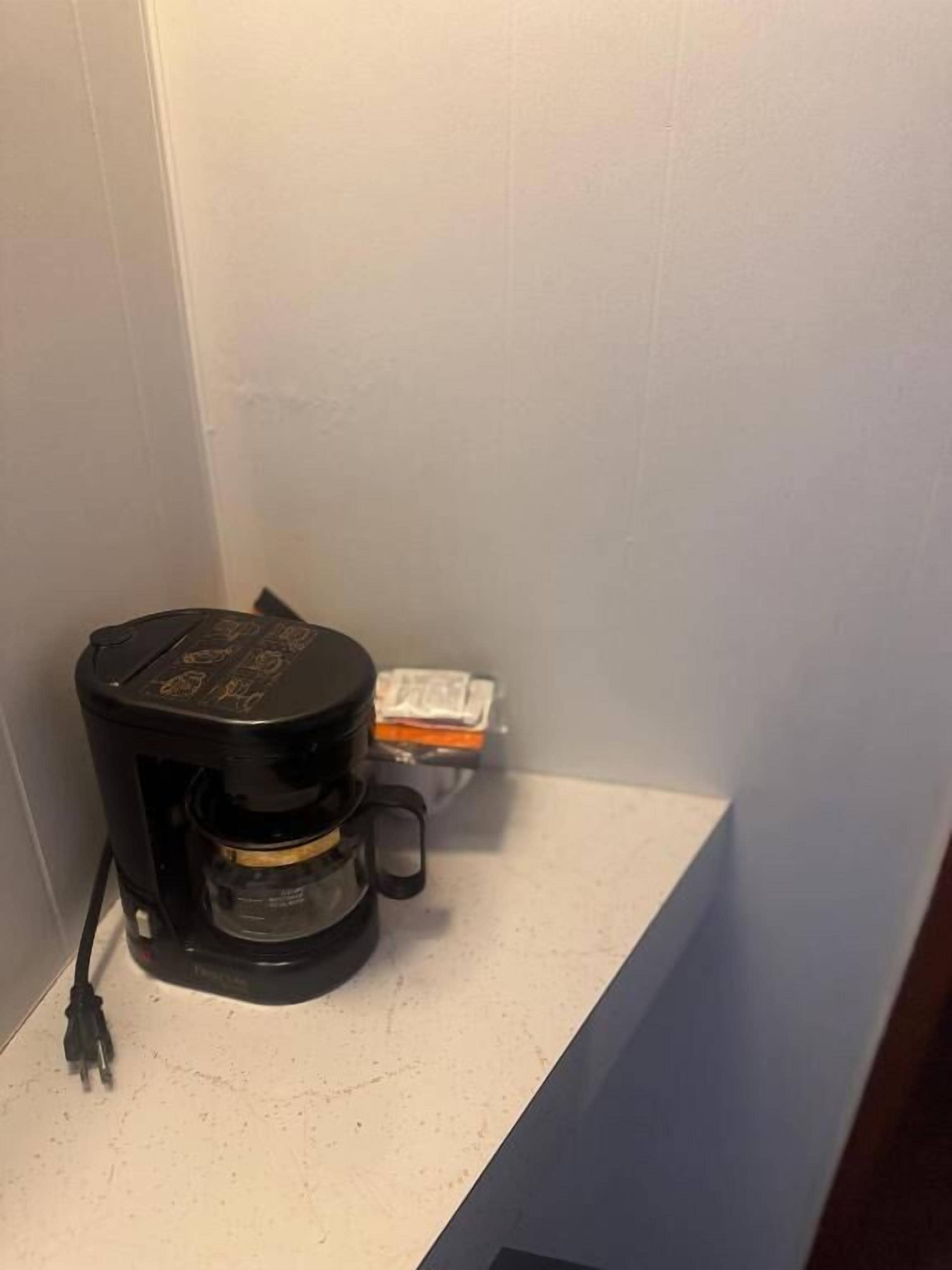 Musta kahvinkeitin valkoisella työtasolla huoneessa, jonka seinät ovat vaalean sävyisiä.