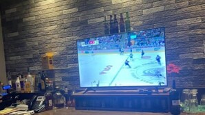 Una zona de bar con una gran pantalla de televisión mostrando un partido de hockey, armarios de madera con etiquetas que indican los tipos de cerveza, y varias botellas y vasos sobre la barra.