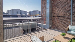 Een terras op het dak, met een bakstenen muur, een metalen hekwerk, een rieten bank, een kleine tafel met een plant, en een uitzicht op gebouwen in de verte.