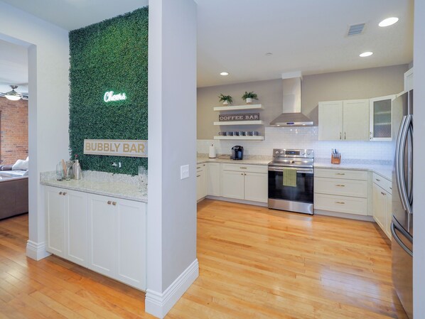 Una cocina moderna con armarios blancos, una pared verde con un letrero que dice 'Coffee Bar', suelo de madera y un refrigerador de acero inoxidable.