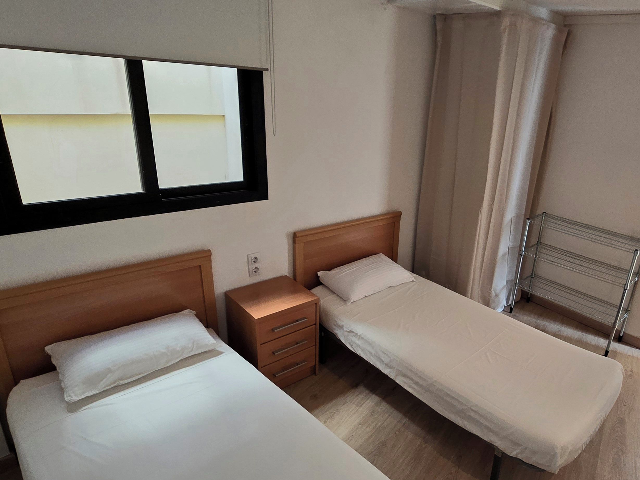 Habitación de hotel con dos camas individuales, una mesita de noche, un pequeño cajón, una ventana con vistas y un estante metálico.