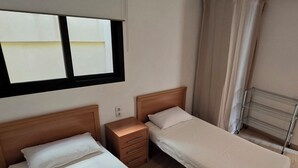 A hotel room with two single beds, a bedside table, a small drawer, a window with a view, and a metal shelf.