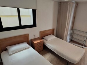 Habitación de hotel con dos camas individuales, una mesita de noche, un pequeño cajón, una ventana con vistas y un estante metálico.