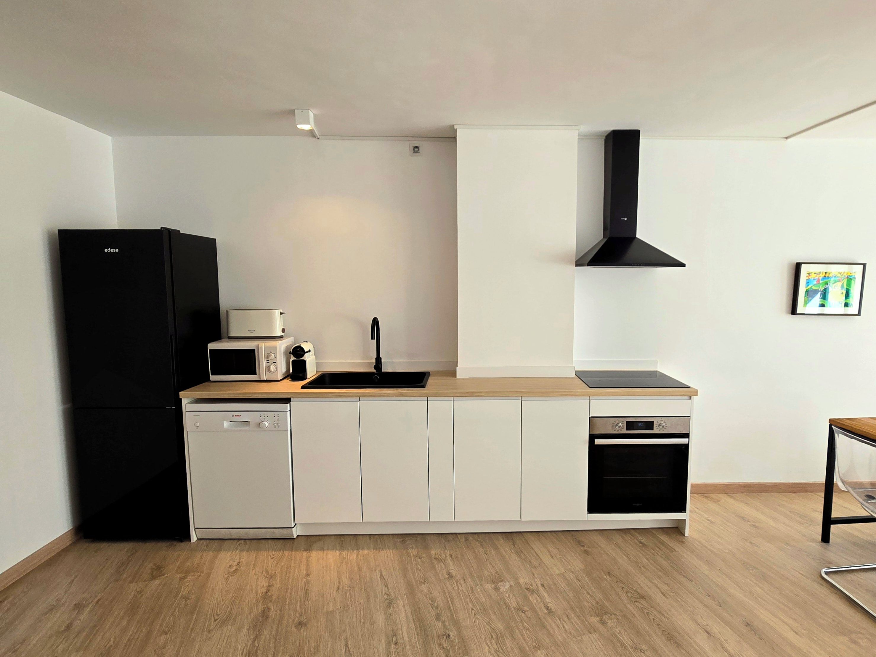 Cocina compacta con refrigerador negro, gabinetes blancos, microondas, tostadora, pileta y campana extractora negra.