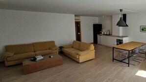 A living room with a sofa, a coffee table, and a dining table with chairs. There is a kitchen area with a refrigerator, microwave, and sink.