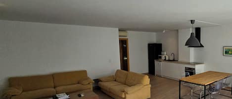 Ein Wohnzimmer mit einer Couch, einem Couchtisch und einem Esstisch mit Stühlen. Es gibt eine Küchenzeile mit Kühlschrank, Mikrowelle und Spüle.
