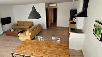 Un espacio habitable moderno con una mesa de comedor de madera, un sofá, un televisor montado en la pared y una zona de cocina.