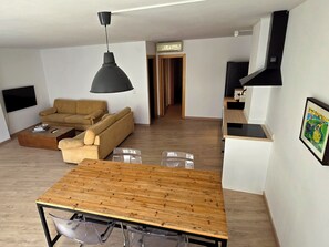 Un espai modern d'estar amb taula de dinar de fusta, sofà, televisió penjada a la paret i zona de cuina.