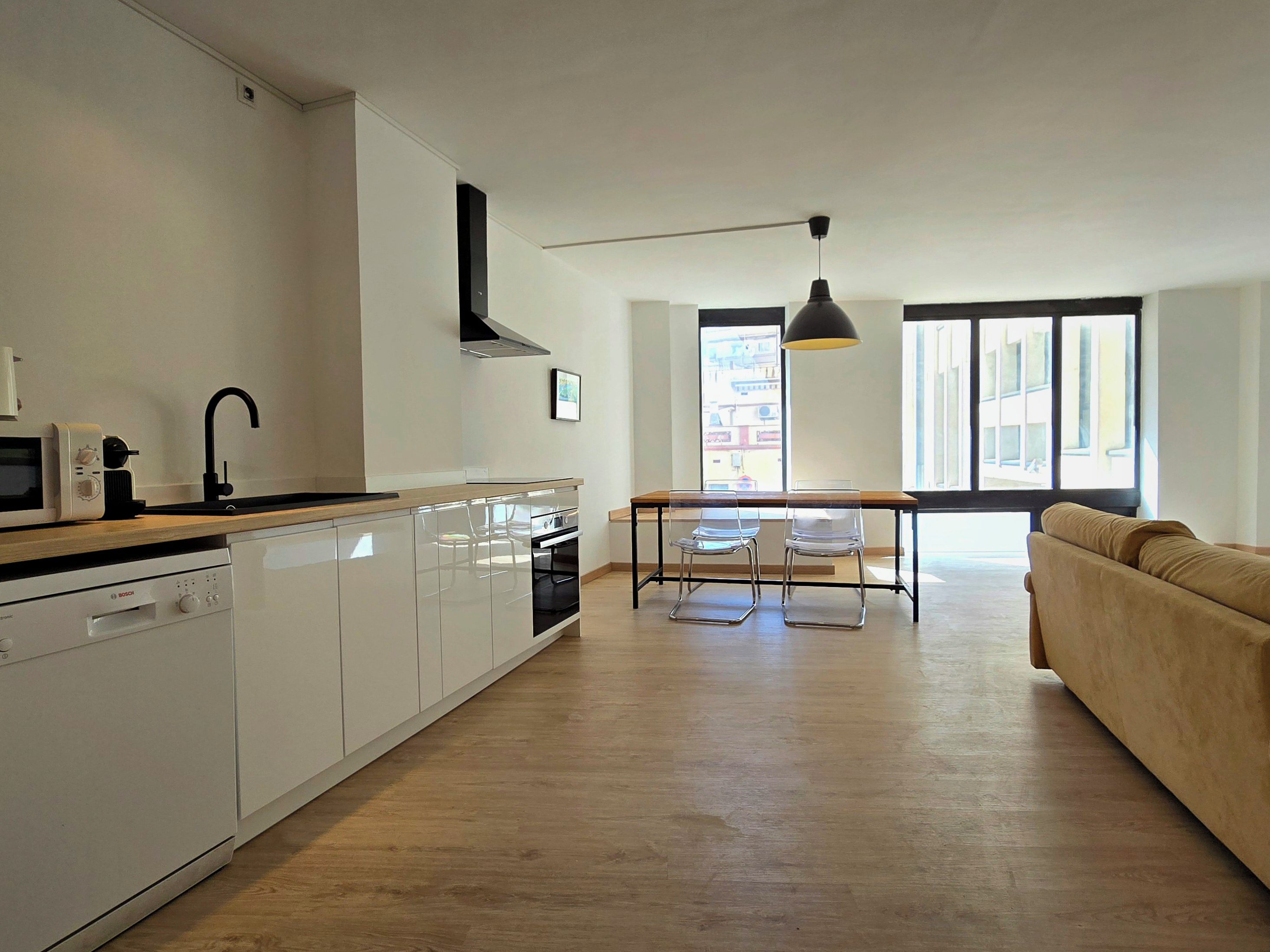 Cocina moderna con armarios blancos, lavavajillas Bosch y grifo negro. Hay una zona de comedor con mesa y sillas, y un sofá en la sala de estar.