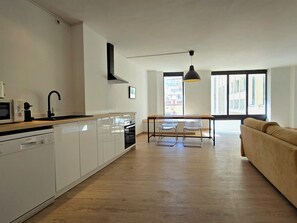 Cocina moderna con armarios blancos, lavavajillas Bosch y grifo negro. Hay una zona de comedor con mesa y sillas, y un sofá en la sala de estar.