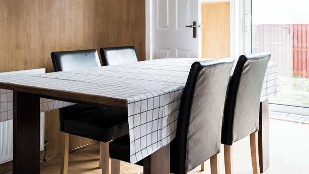 Een eetruimte met een lange tafel, vier stoelen, en zicht op een houten deur en een rode hek buiten.