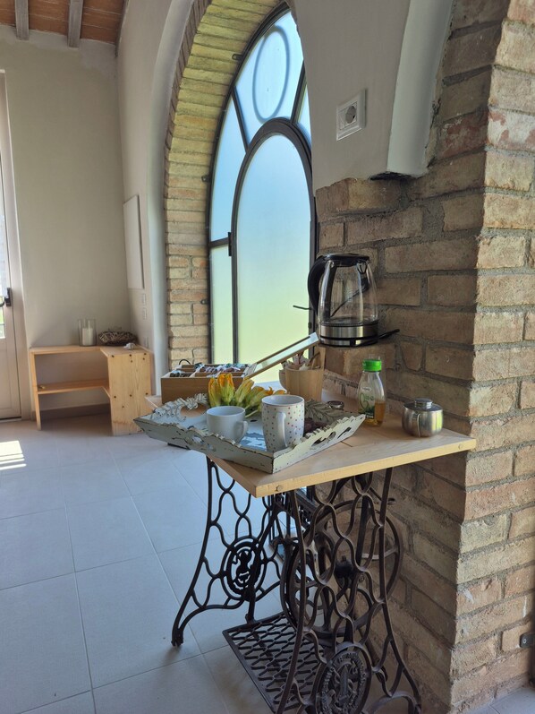 A cozy room with a wooden table set with a tray, a coffee pot, and various items. A window with a unique design is visible in the background.