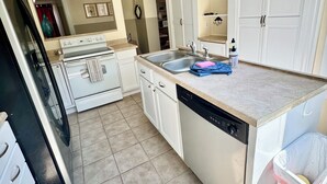 Una cocina con armarios blancos, lavavajillas, estufa y lavabo. Hay un cubo de basura y una lámpara en el techo.