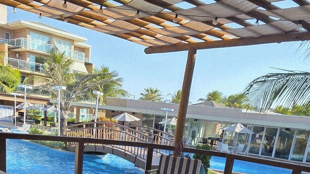 A poolside area with wooden pergolas, lounge chairs, and a view of a building in the background.