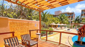 A covered outdoor seating area with wooden furniture, a pool, and a view of a residential building.