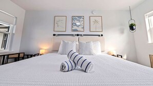 Ein ordentlich bezogenes Bett mit einem gestreiften Handtuch, ein Kopfteil mit einer horizontalen Leiste und drei gerahmte Kunstwerke an der Wand.