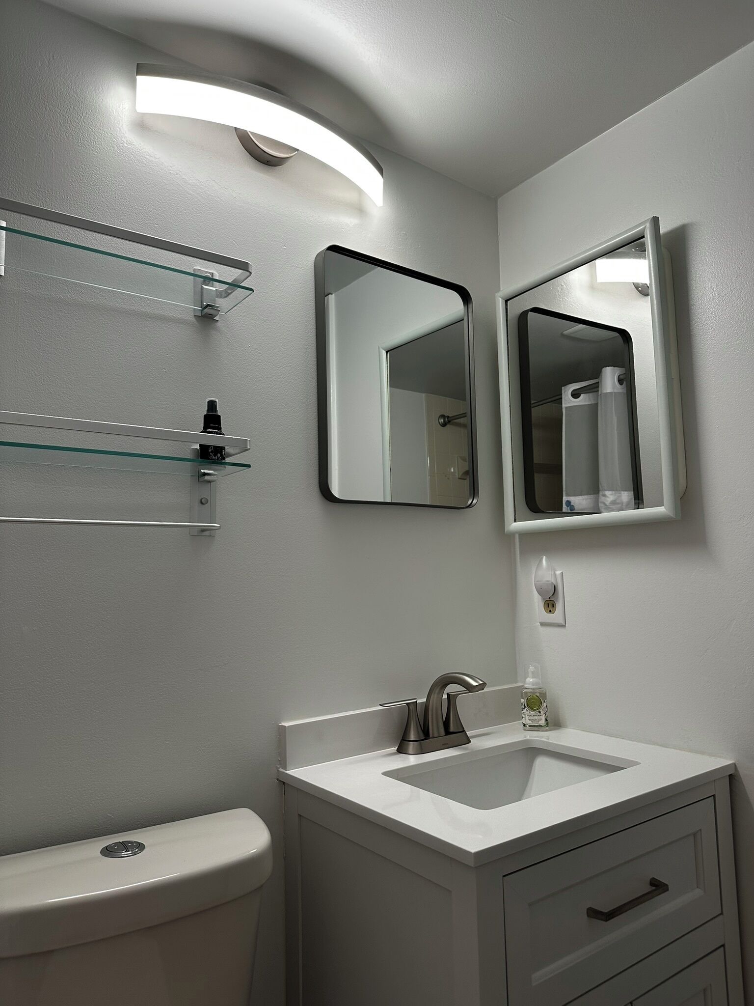 A modern bathroom with a white sink, a toilet, and two mirrors. There are glass shelves above the sink and a shower curtain visible in the mirror reflection.