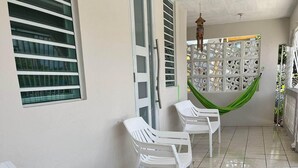 A clean, tiled patio area with white plastic chairs and a table, a door with a window, and a decorative wall with a hanging hammock. - Vieques Beach Home, Boho style, within walking distance to beach and a King Bed (Florida)